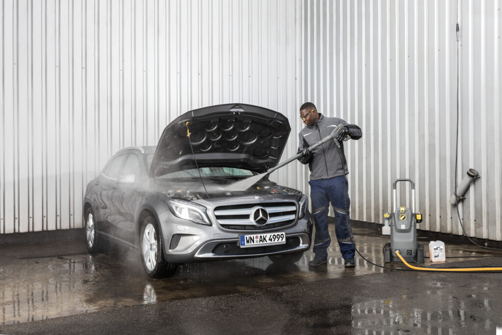 Mężczyzna w szarej kurtce myje urządzeniem ciśnieniowym silnik w samochodzie marki Mercedes
